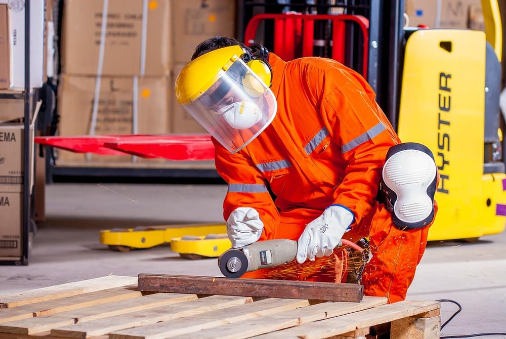 A worker in orange hi-vis coveralls, full face shield, ear defenders and gloves cutting steel with an angle grinder beside a forklift and stacked pallets.