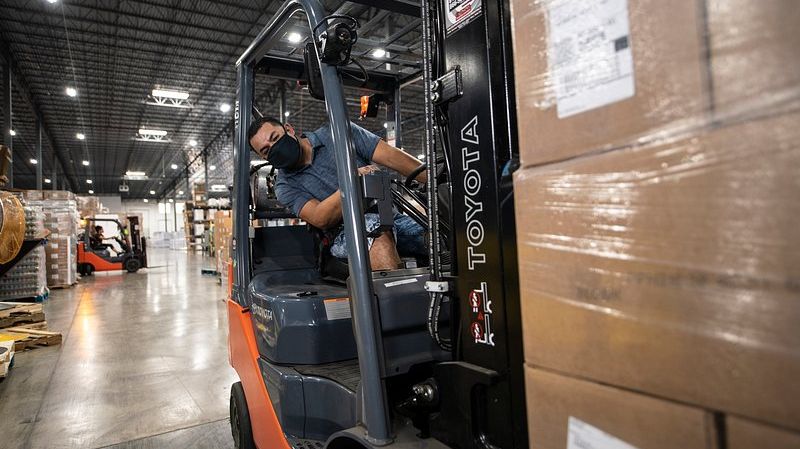 A forklift operator moving a shrink-wrapped pallet down a warehouse aisle.